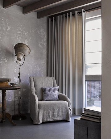Cozy corner with a gray armchair, standing lamp, side table, and window dressed in fabric blinds. Books are stacked on a nearby surface.