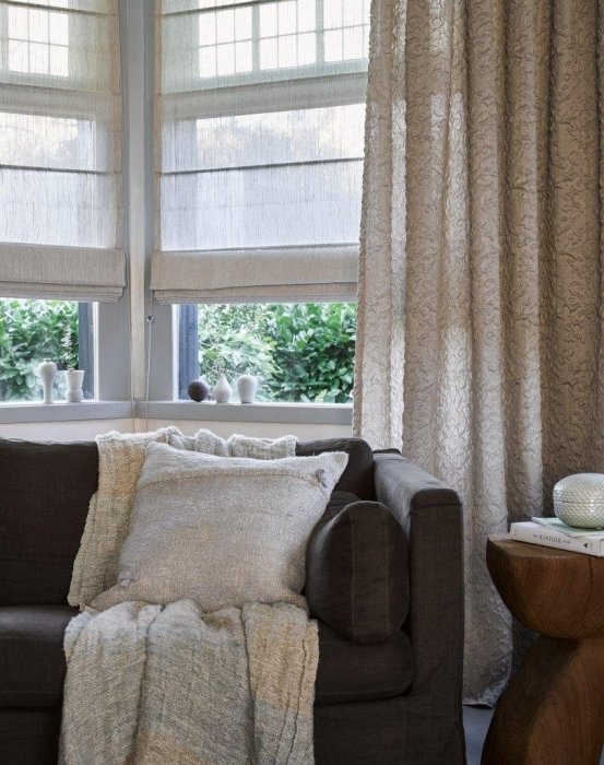 A cozy living room corner features a dark sofa adorned with a light throw blanket and pillows. Sheer blinds gently filter the sunlight through the windows, while a small wooden side table with a candle completes this soothing space.