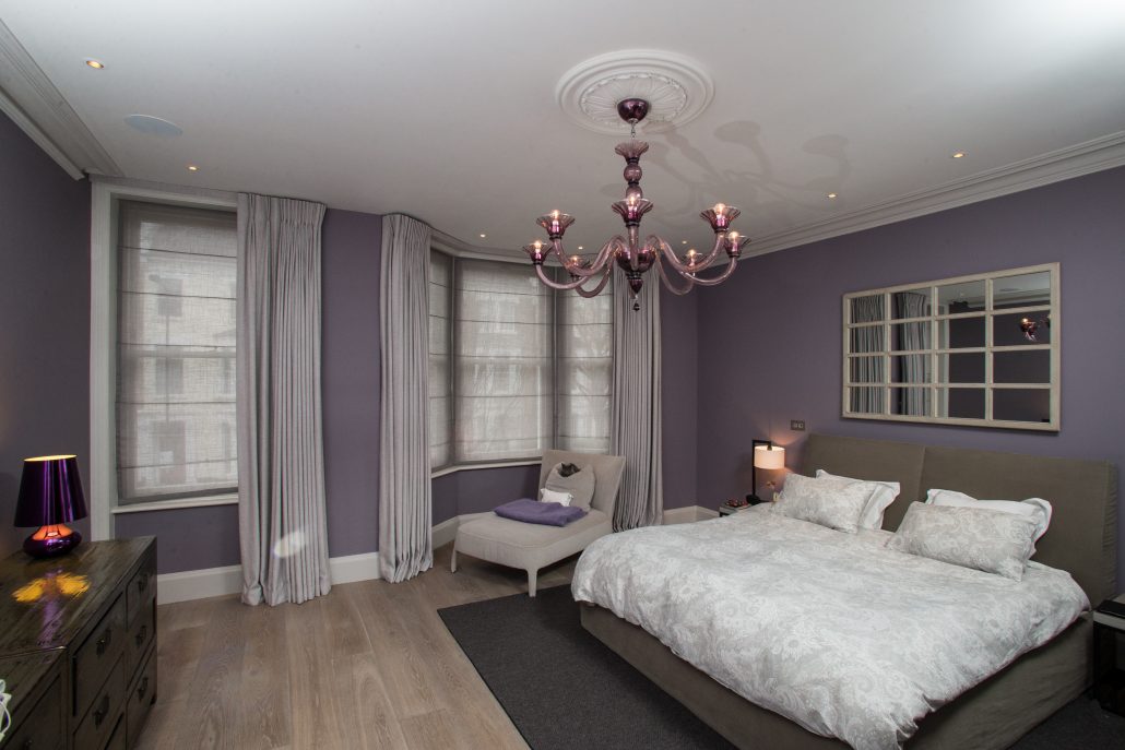 A bedroom with lilac walls features a bed, chair, and dresser beneath a purple chandelier. Two windows on the left wall are adorned with colorful Roman Blind Range A, adding vibrant charm to the serene space.