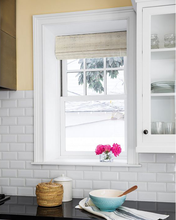 The kitchen window, framed in white with a beige fabric blind, offers a serene view of the tree outside. A bowl with a whisk sits on the countertop alongside a basket and pink flowers in a vase. The backsplash is tiled in white, complementing the cabinet filled with glasses.