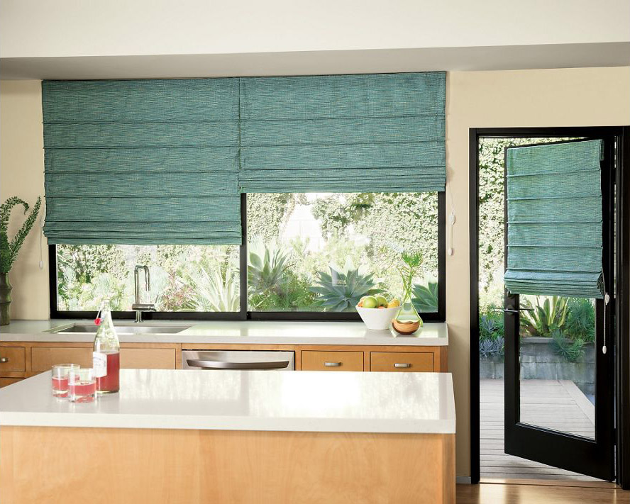 Kitchen with a white countertop, wooden cabinets, and large windows adorned with teal fabric blinds. A glass door opens to a leafy garden, while a vibrant bowl of fruit sits on the counter.