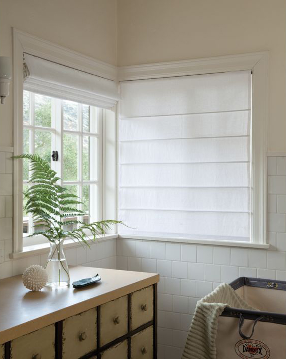 Bright corner room with white Roman shades and blinds on the windows. A fern in a glass vase sits on the countertop alongside a small decorative object. Cream-colored tiles and a crib are partially visible, adding a touch of calming colour to the space.