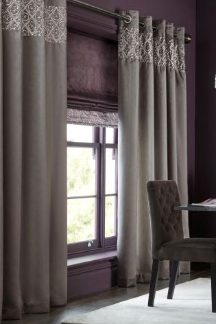The room features purple walls with a window bathed in light. Gray curtains adorned with patterned tops hang gracefully, complemented by a dark upholstered chair. The subtle play of colour highlights the exquisite fabric textures throughout the space.