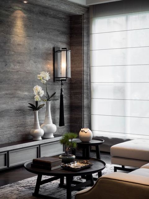 A modern living room features a stunning marble wall and a sleek, wall-mounted light fixture. White orchids in vases complement two round black tables with decor, while a white sofa faces the large window adorned with sheer fabric blinds.