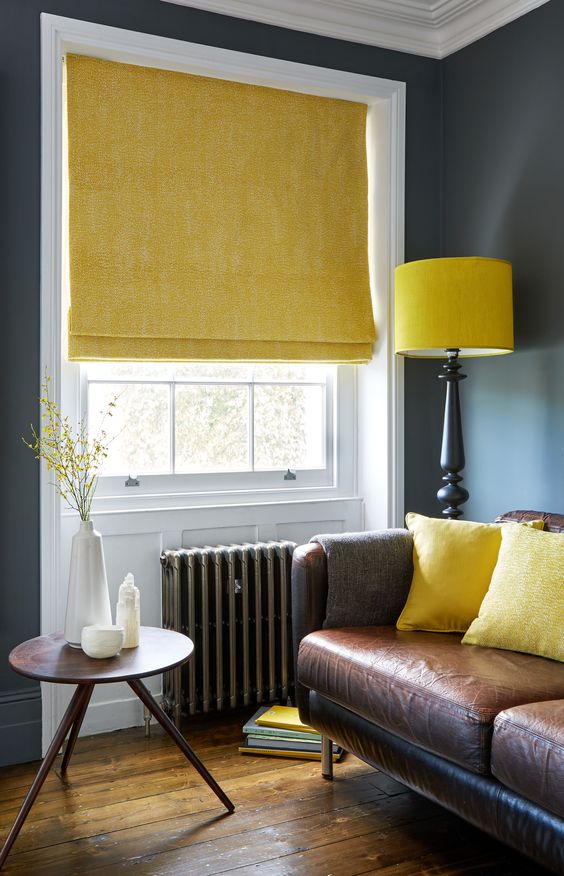The living room features a leather sofa adorned with yellow cushions, complemented by a matching yellow blind and lamp. A small round table sits near the window, offering views of lush greenery. The muted teal walls perfectly harmonize the colors and fabrics in this inviting space.