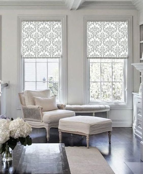 Elegant living room with patterned roller blinds in a rich fabric, a cushioned armchair, ottoman, and chaise lounge. Features a white floral arrangement and hardwood floors that complement the vibrant colours.