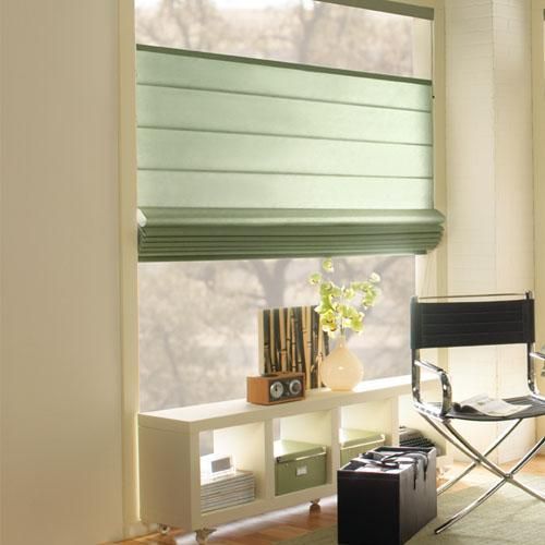 A room with green Roman shades and fabric blinds, a black chair, and a white shelf holding decorative items and a plant. Soft natural light filters through the window.