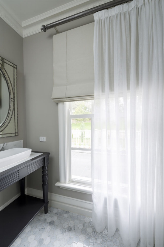 A bathroom with a tall window featuring sheer white curtains and a gray fabric Roman shade. There is a black vanity with a white sink next to the window, subtly complemented by elegant blinds that add privacy and style.