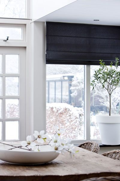 A room with a wooden table, vibrant white flowers in a vase, and a potted plant near large windows adorned with sleek black blinds. The snowy scene outside offers a stunning contrast of colour against the serene interior.