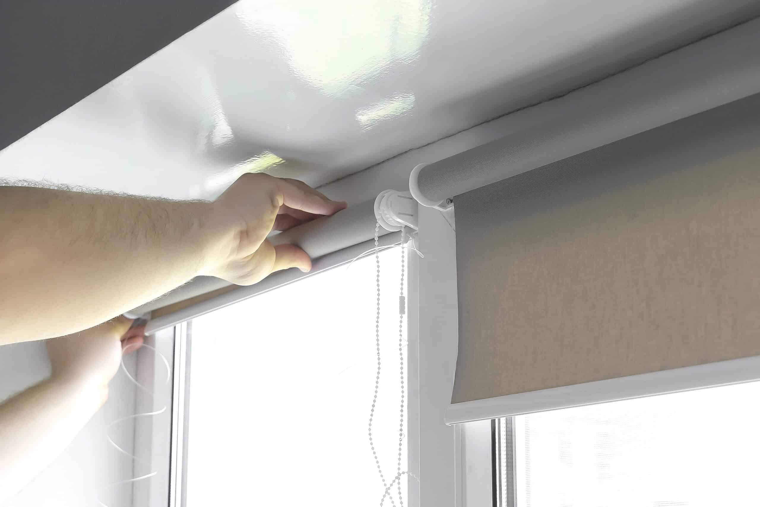 Hands adjusting a gray roller blind on a window with a white wall background.