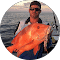 A person holds a large, vibrant orange fish against the backdrop of the ocean, its colour as striking as vibrant fabric swaying in the gentle breeze.