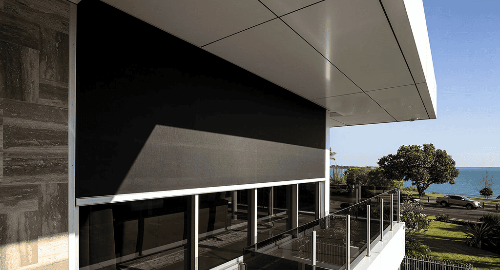 A modern home showcases a large black Outdoor Straight Drop Blind on the upper balcony, complemented by glass railings and scenic views of trees and water in the background.