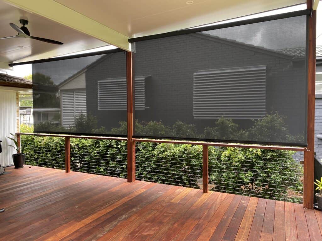 A covered wooden deck with cable railing features Harbour Zebra Blinds facing a house with horizontal shutters. The ceiling includes a fan and recessed lighting.