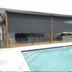 A covered outdoor patio features two large, partially lowered grey Outdoor Straight Drop Blinds beside a swimming pool with clear water and tile edging.