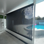 An Outdoor Straight Drop Blind in black is partially lowered, providing shade to a covered patio area beside a backyard pool.
