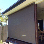 An Outdoor Straight Drop Blind in brown is fully lowered, providing complete coverage for a patio or verandah area attached to a house.