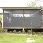 A house with a screened-in porch elevated on wooden posts features Outdoor Straight Drop Blinds with black mesh screens, complemented by small green bushes planted along the base.