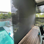 Modern backyard with an Outdoor Straight Drop Blind and wooden flooring on the balcony, featuring a dark privacy screen overlooking the pool, patio furniture, and garden area.