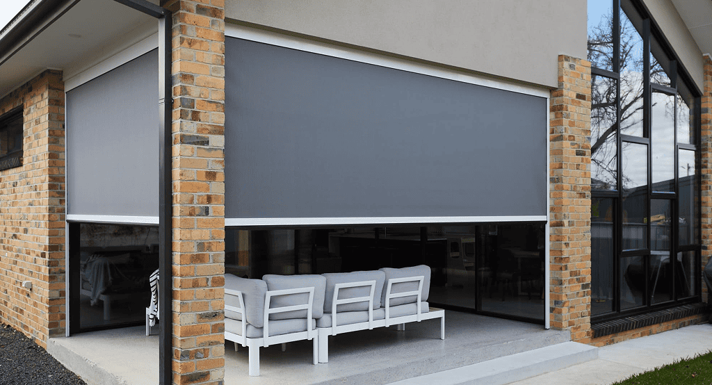 A covered outdoor patio with a brick exterior features large gray Outdoor Straight Drop Blinds and white patio furniture with gray cushions.