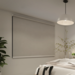 A modern bedroom is styled with Premium Range Roller Blinds, a white pendant light, a leafy potted plant, and minimal decor on the side table.