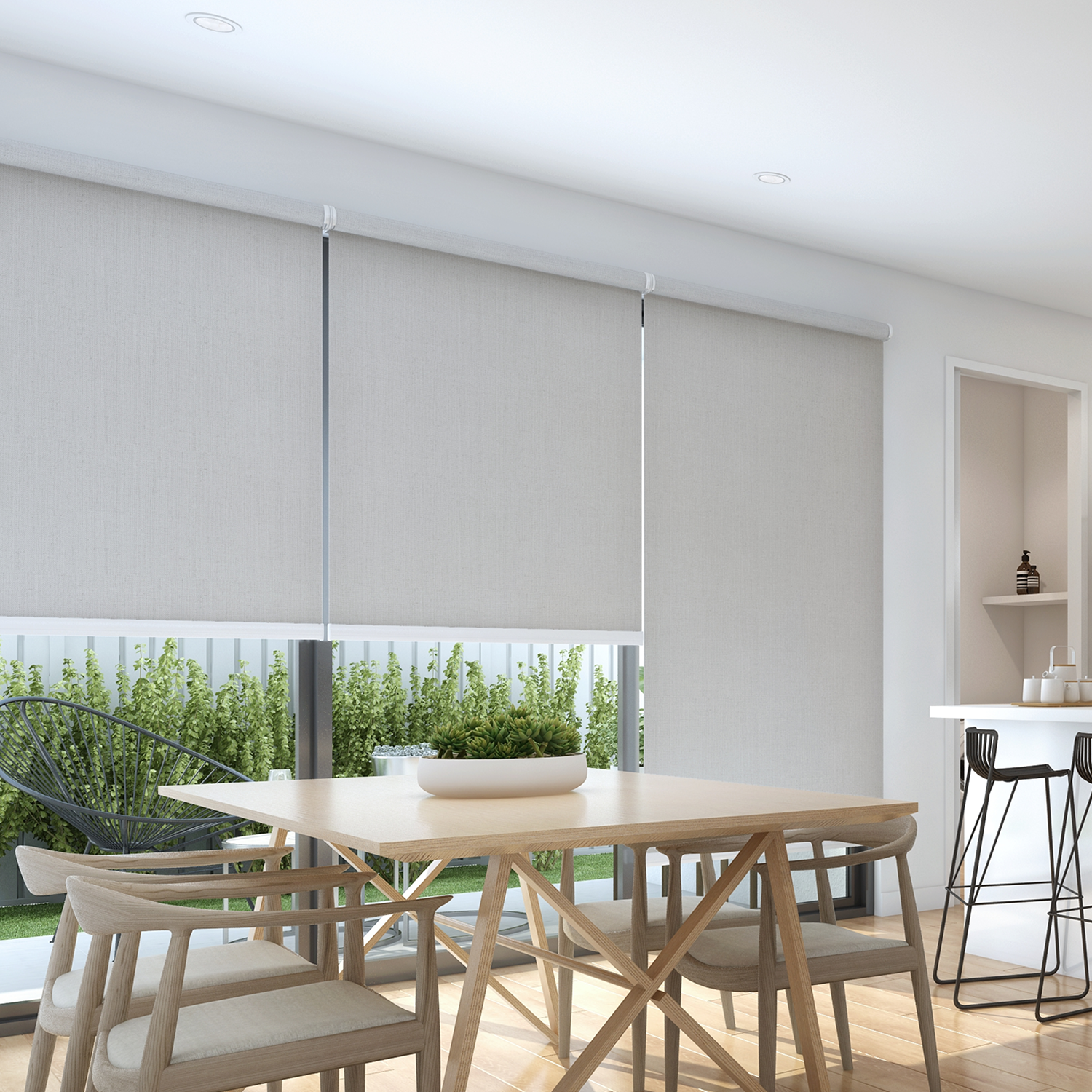 A stylish dining area features a wooden table and chairs by a large window fitted with Premium Range Roller Blinds, offering views of lush greenery outside.
