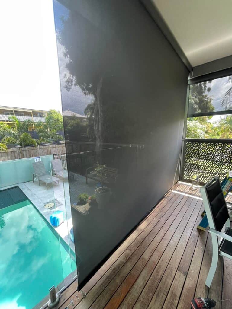 A Harbour Zebra Blind is partially rolled down on a wooden deck overlooking a swimming pool and garden area.