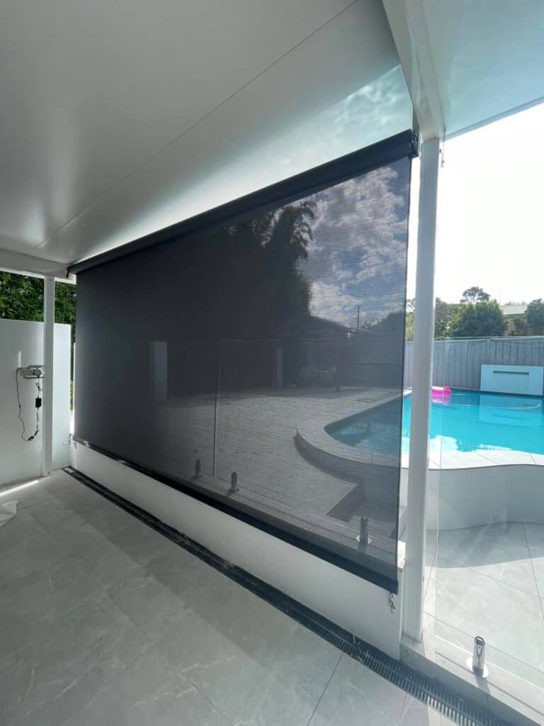 The Harbour Zebra Blind in black partially covers a white poolside patio, with a pool and trees visible in the background.