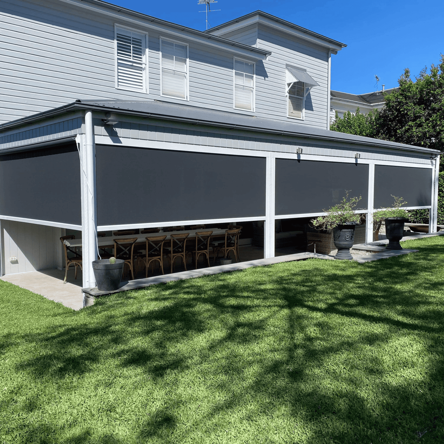 A covered patio features Outdoor Deep Channel Blinds, wooden dining chairs, potted plants, and overlooks a large green lawn in front of a two-story house.