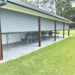A covered outdoor patio with a row of chairs and tables is enclosed by large gray Outdoor Deep Channel Blinds, next to a lawn.