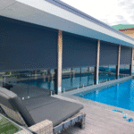 A modern poolside area features a lounge chair, wooden deck, glass fence, and large Outdoor Deep Channel Blinds covering the windows beside the pool.