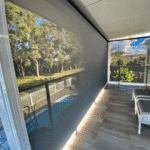 An Outdoor Deep Channel Blind is partially rolled down on a covered patio, reflecting nearby trees and a fenced pool area in the background.