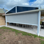 A covered outdoor patio features Outdoor Deep Channel Blinds, with a large, rolled-down grey screen shading a dining table and chairs under a peaked roof.