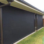 A house with two large Outdoor Deep Channel Blinds covering the windows, beside a lawn and a hose on the ground.