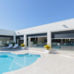 Modern white house with large windows, featuring a swimming pool and patio with lounge chairs in the foreground, set against a clear blue sky.