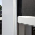 Close-up of a modern window corner with a mesh screen, featuring sleek white frames. Background shows rooftops and trees against a clear sky.