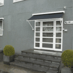A modern glass double door features Outdoor Pivot Arm Blinds above, set in a gray building with tiled steps and two square planters holding trimmed bushes on either side of the entrance.