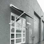 Outdoor Pivot Arm Blinds are installed over a glass door on the exterior of a commercial gray building, positioned next to a closed garage door.