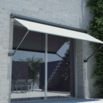 White Outdoor Pivot Arm Blinds extend over a large glass sliding door set in a stone wall, reflecting outdoor stairs and patio furniture.