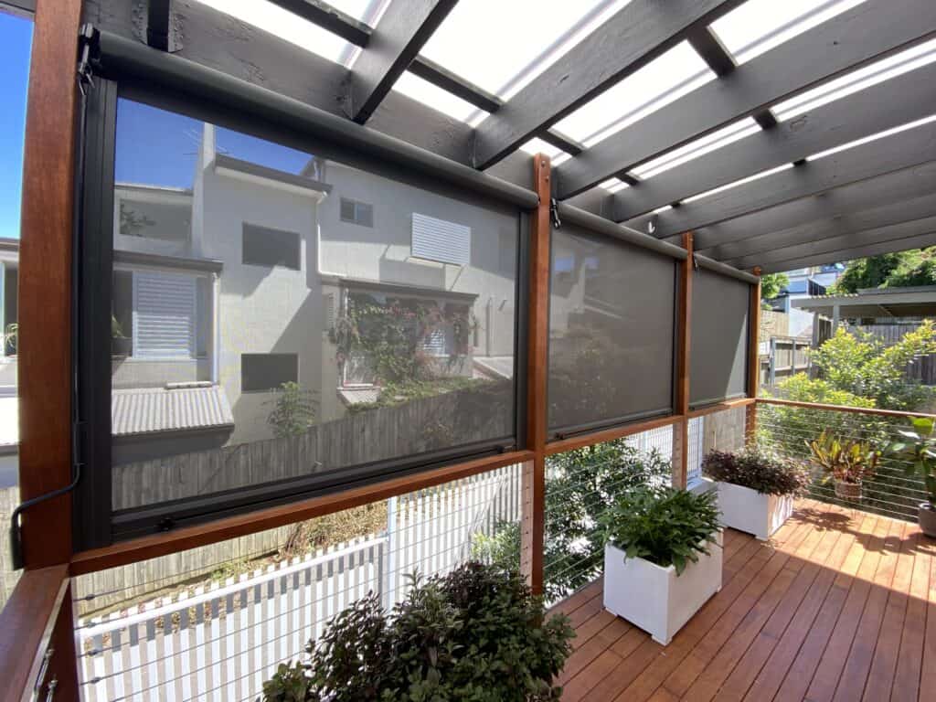 Outdoor covered patio with wooden flooring, potted plants, and retractable mesh blinds partially rolled down, overlooking neighboring houses and a fenced yard.