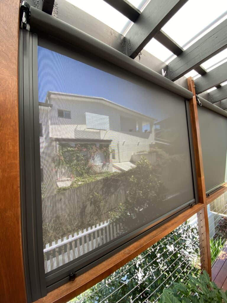 A mesh outdoor roller blind partially lowered on a patio, with a house, garden, and white fence visible through the screen.