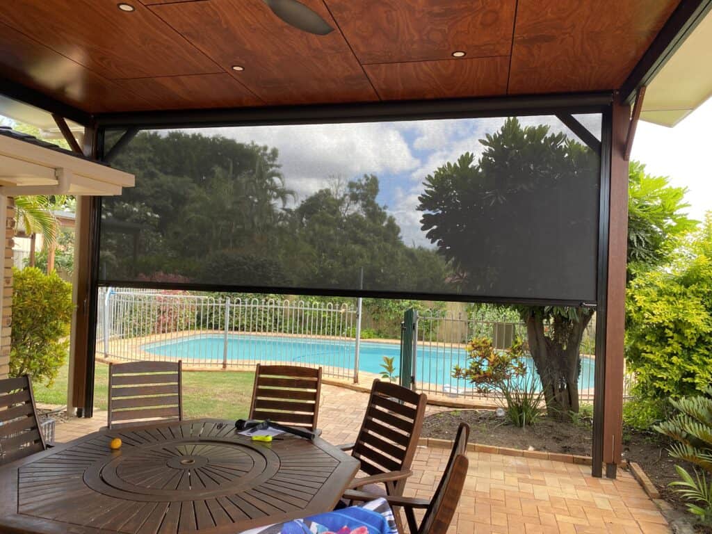 Covered outdoor patio with a round table and chairs, overlooking a fenced backyard swimming pool and garden, with a mesh sunshade partially lowered.