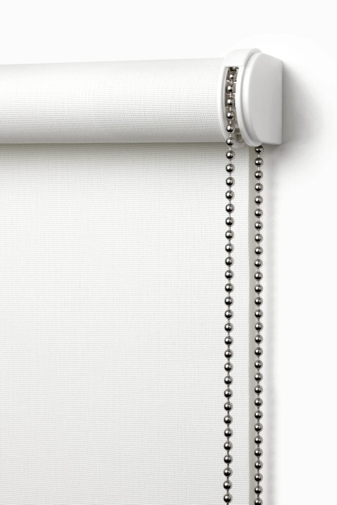 A white roller blind with a metal bead chain hangs against a wall.
