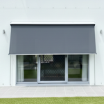A modern grey retractable awning extends over sliding glass doors with white curtains and Pivot Arm Blinds, overlooking a tiled patio and green lawn.