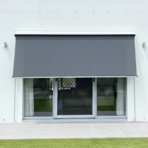 A modern grey retractable awning extends over sliding glass doors with white curtains and Pivot Arm Blinds, overlooking a tiled patio and green lawn.