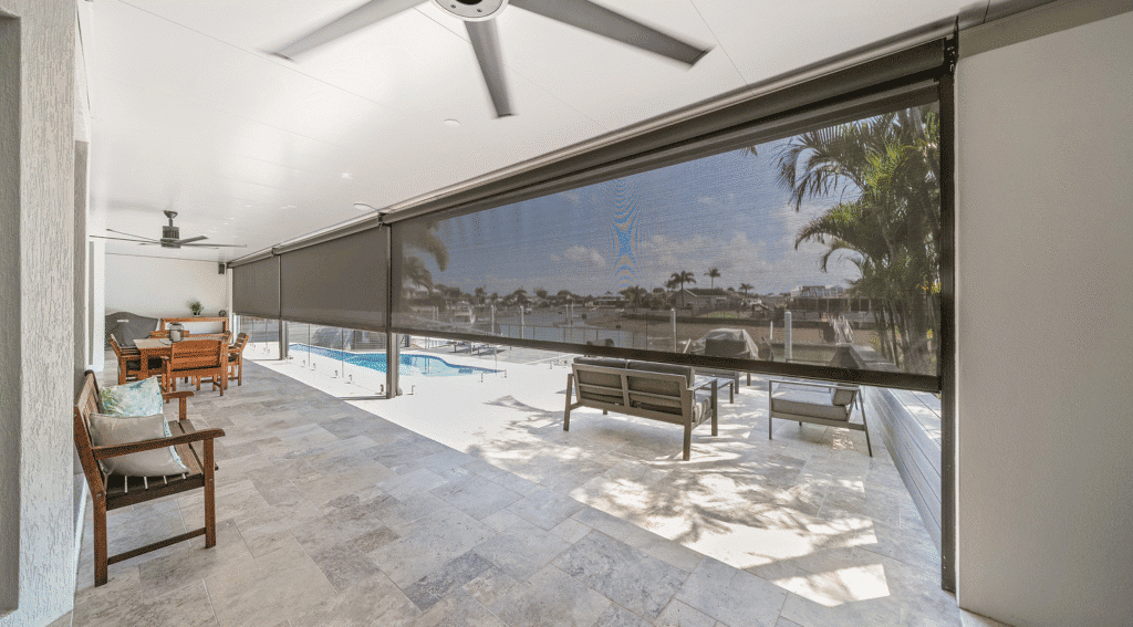 A covered patio with ceiling fans, outdoor seating, a dining area, and retractable mesh screens overlooks the pool and waterfront—upgraded with Outdoor Zipscreen Blinds for added comfort and contemporary style.