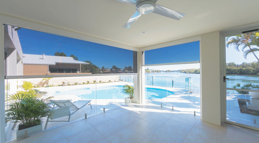 A spacious covered patio with ceiling fan and Outdoor Zipscreen Blinds overlooks a private pool with a glass fence, offering tranquil river and tree views under clear blue skies.
