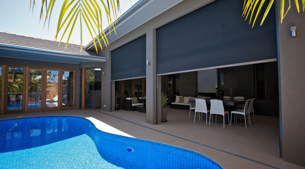 Modern patio with pool, outdoor dining set, and lounge seating, partially enclosed by Outdoor Zipscreen Blinds; palm leaves seen in the foreground.