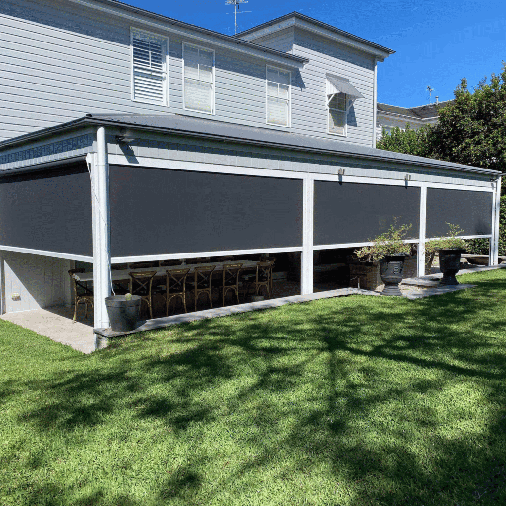 Outdoor patio area with black retractable blinds covering the sides, wooden dining table and chairs underneath, and large potted plants along the perimeter.