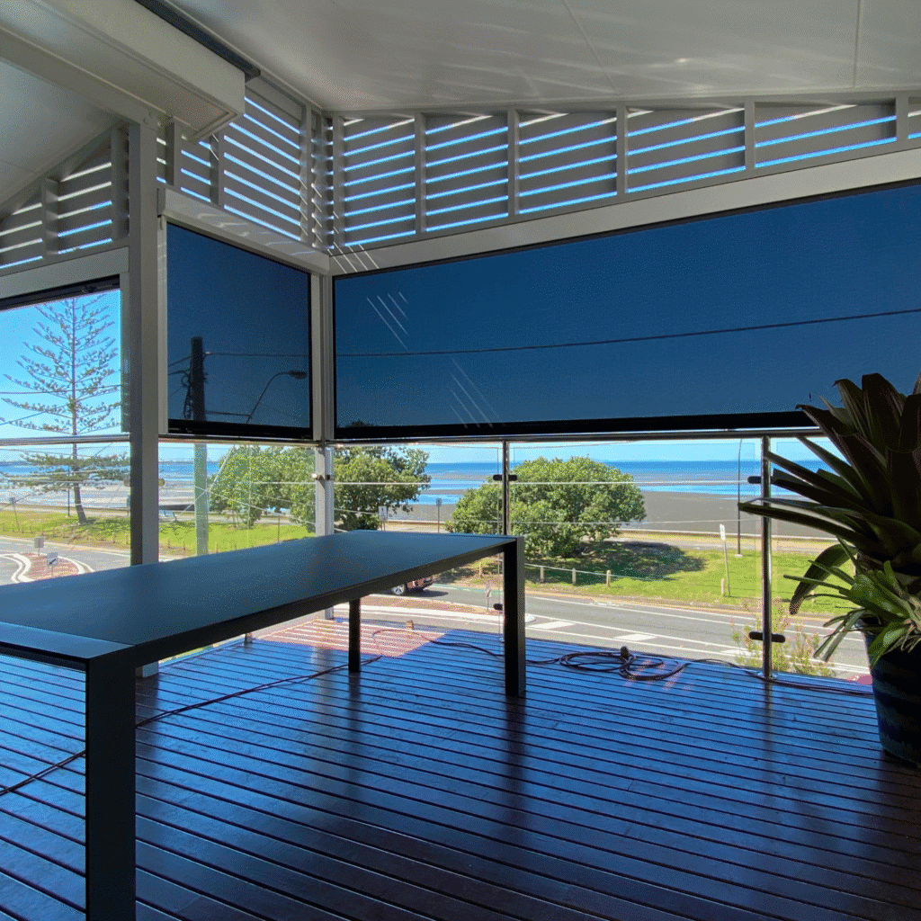 Modern balcony with a glass railing, wooden floor, table, and potted plant, overlooking a street, trees, and a sandy beach in the background under clear blue skies.