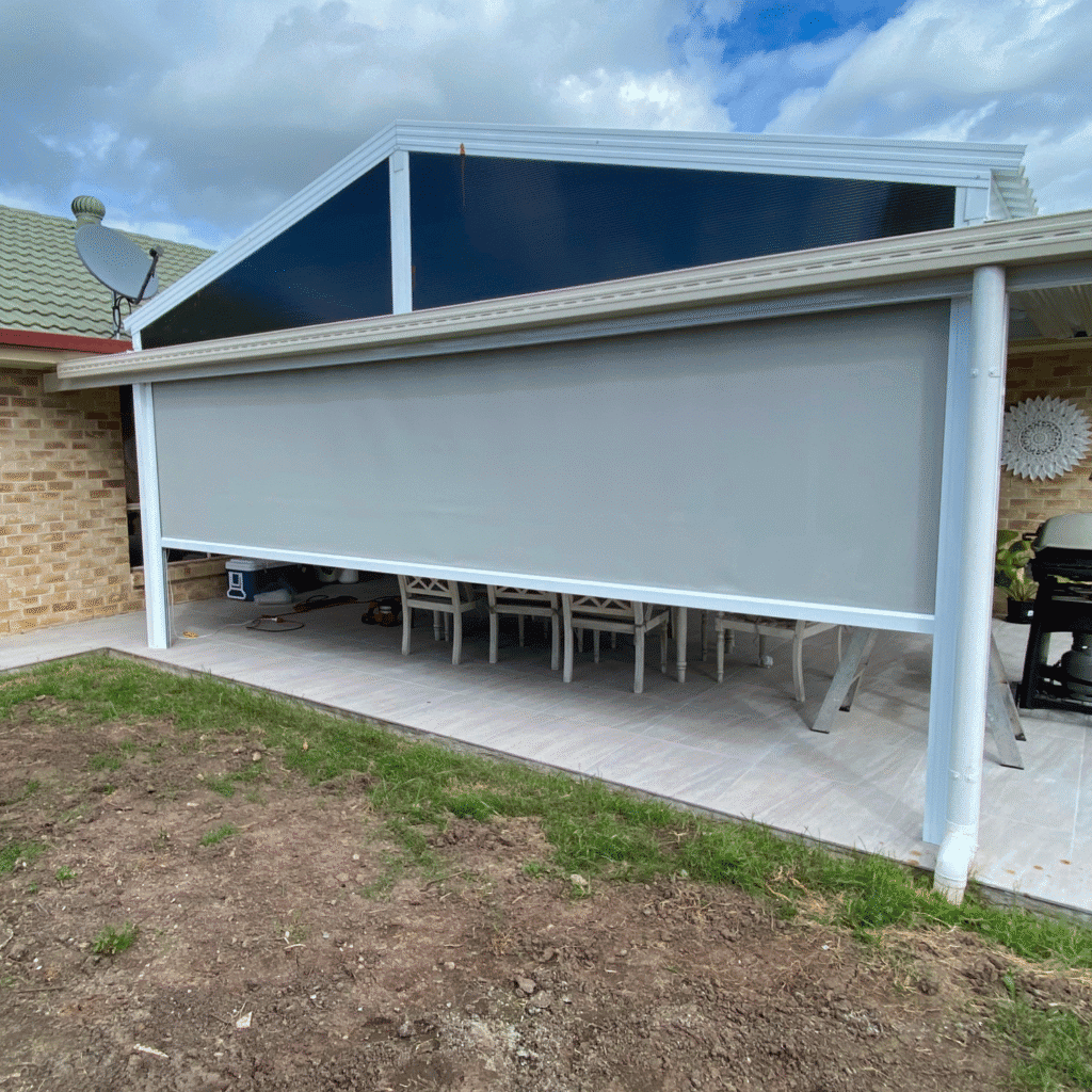 A covered outdoor patio with a lowered grey roller shade, white chairs around a table, and a barbecue grill to the right.